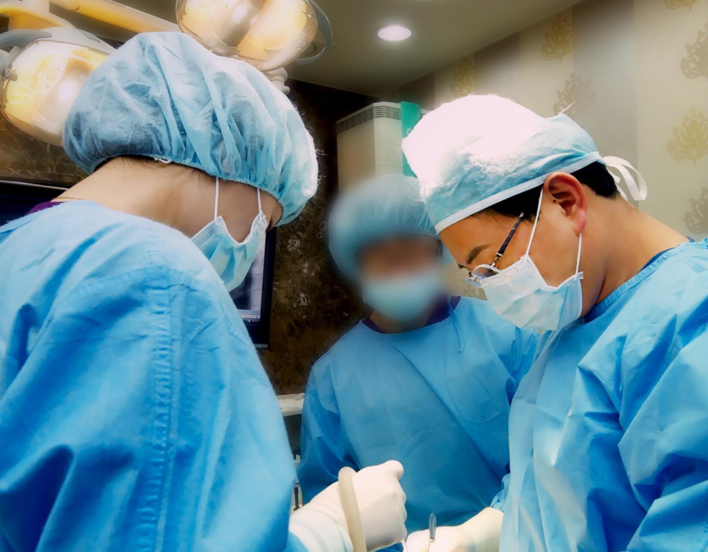 Three dental surgeons in blue scrubs, caps and masks performing a procedure under overhead surgical lights in clinic room.