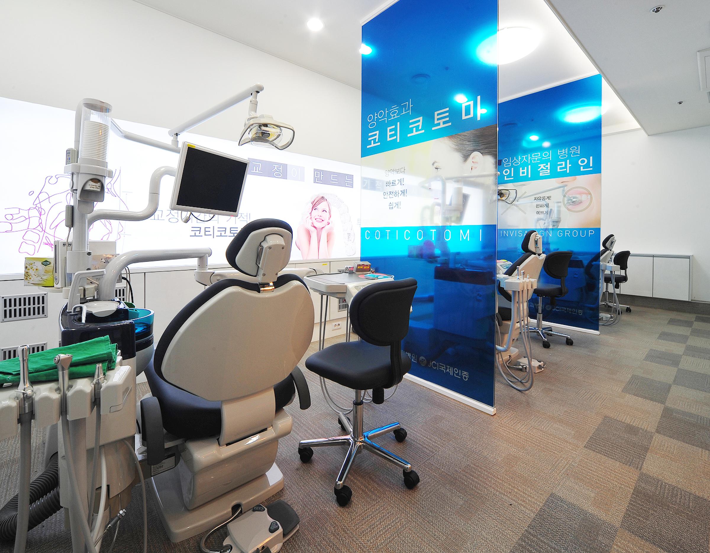 Dental operatory with reclining patient chair, monitor, overhead light and blue privacy panels at 굿윌치과병원 | 부산 JCI 국제인증 치과