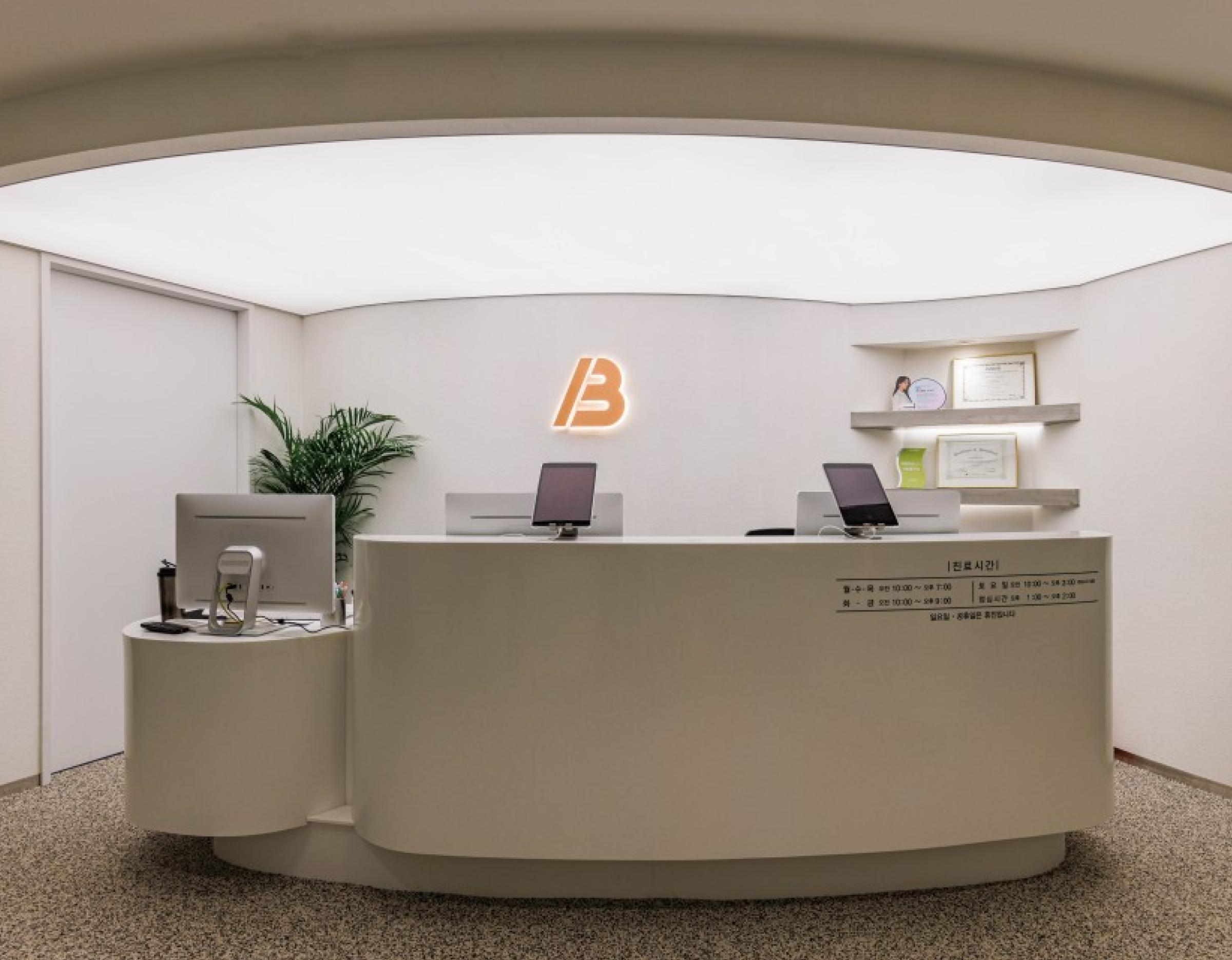 Minimalist curved white reception desk with two laptops, monitors, illuminated "B" logo on wall and potted plant behind it