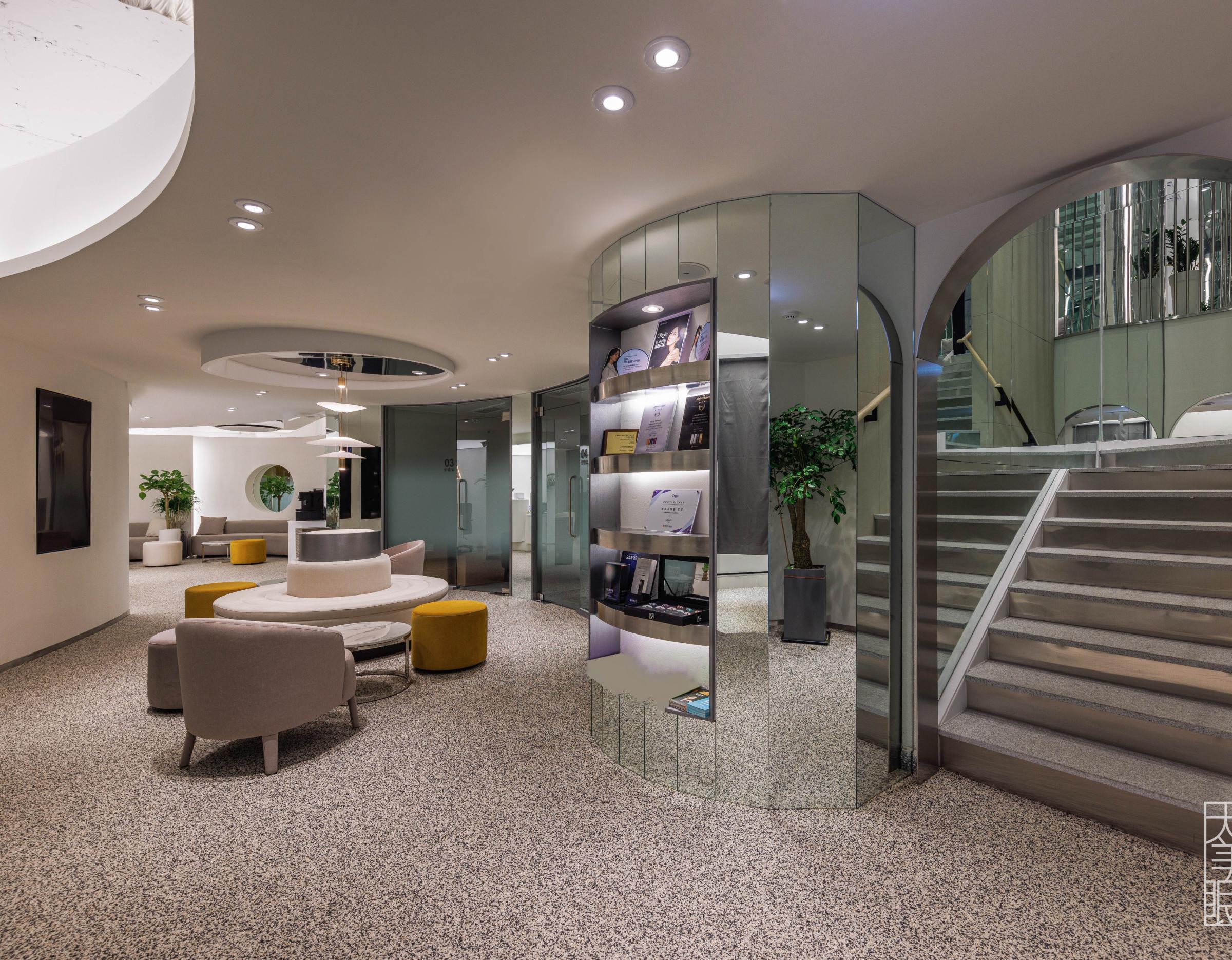 Modern clinic waiting area with circular seating, yellow ottomans, mirrored display column and staircase to upper floor.
