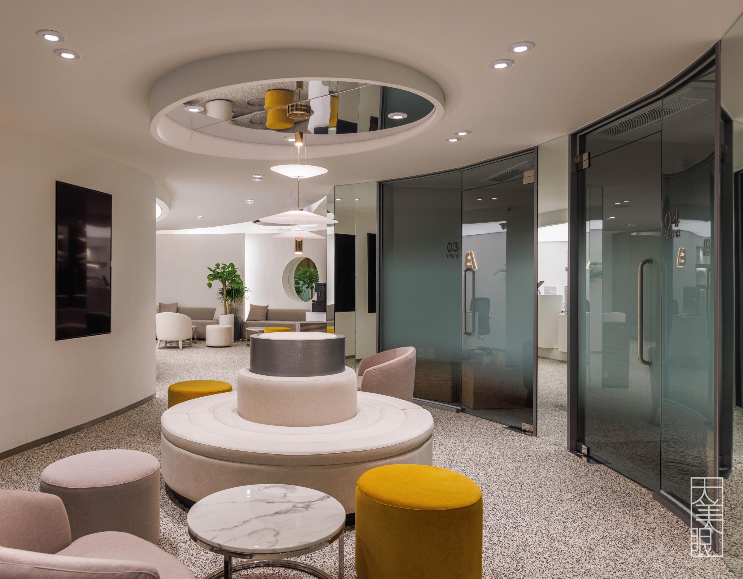 Modern clinic waiting area with circular tiered seating, mustard and pink ottomans, marble side tables and glass consultation rooms.