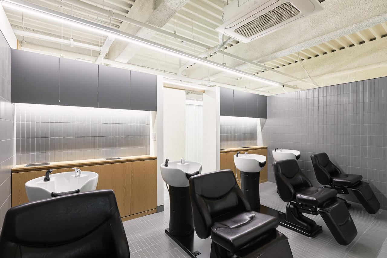 Minimalist salon wash area with three reclining black leather shampoo chairs and white washbasins against grey tiles