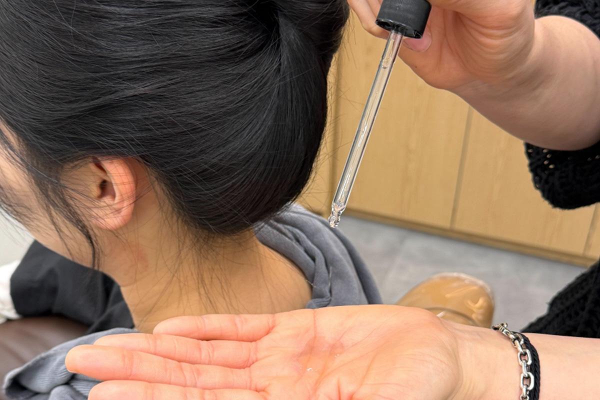 therapist dropping clear scalp tonic from a glass pipette onto palm next to client's tied-back dark hair and neck