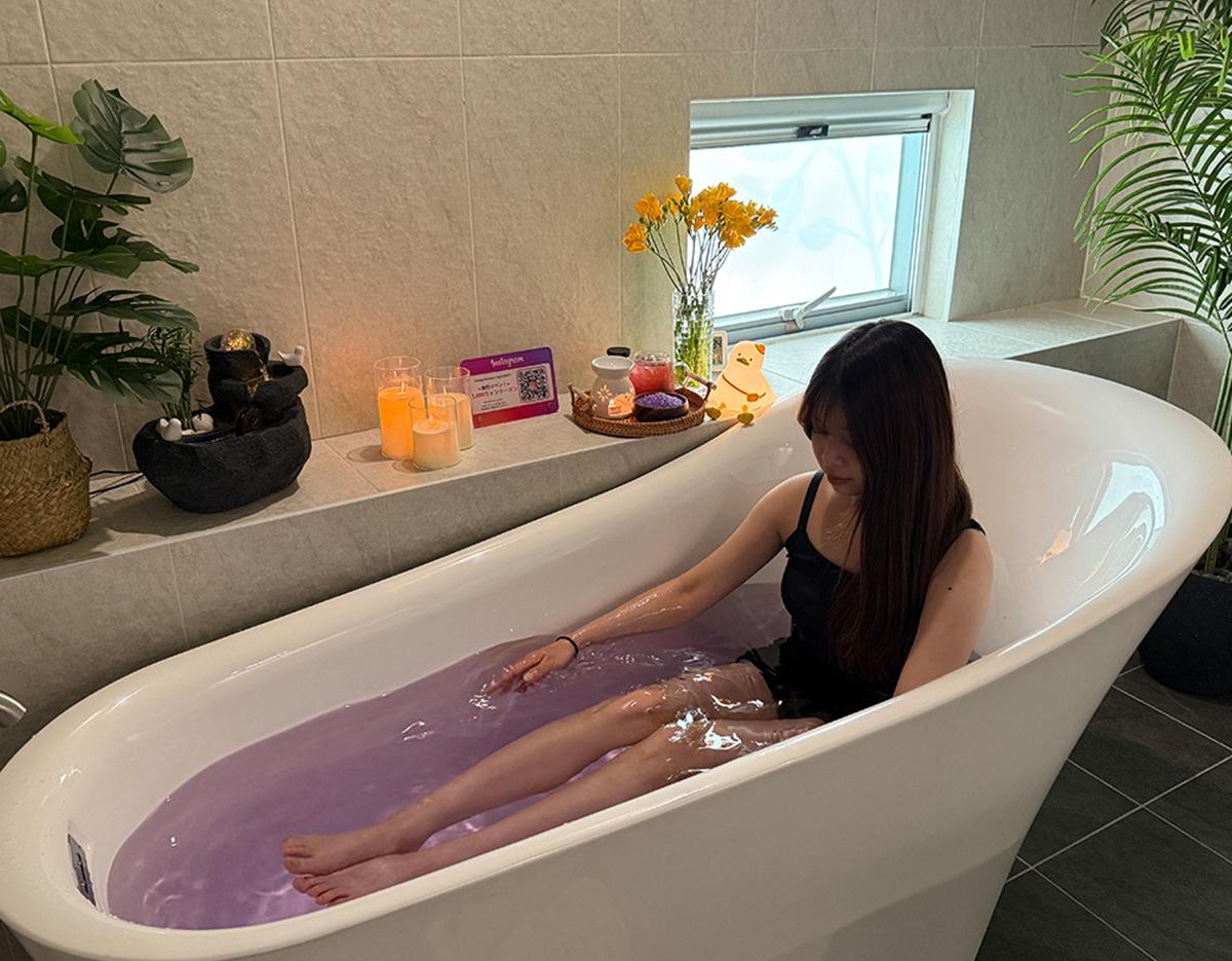 woman in black swimwear reclining in a white freestanding tub filled with lavender-colored water, candles and plants nearby