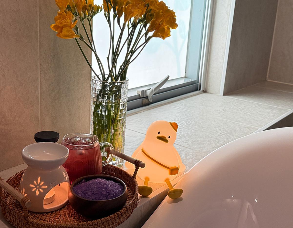 bathside tray with oil burner, bowl of purple bath salts, iced drink, vase of yellow flowers and a small duck-shaped night light