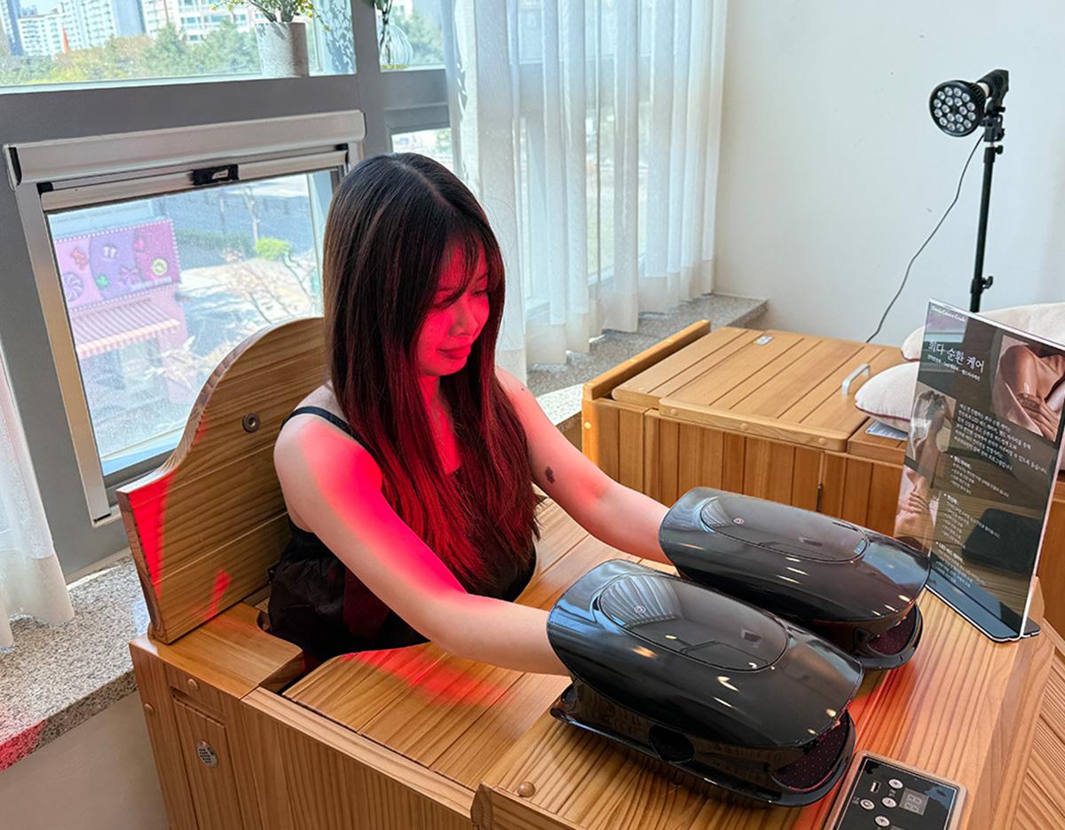 Woman with face obscured sitting in wooden spa chair, hands inside two black hand massage devices under red therapy light.