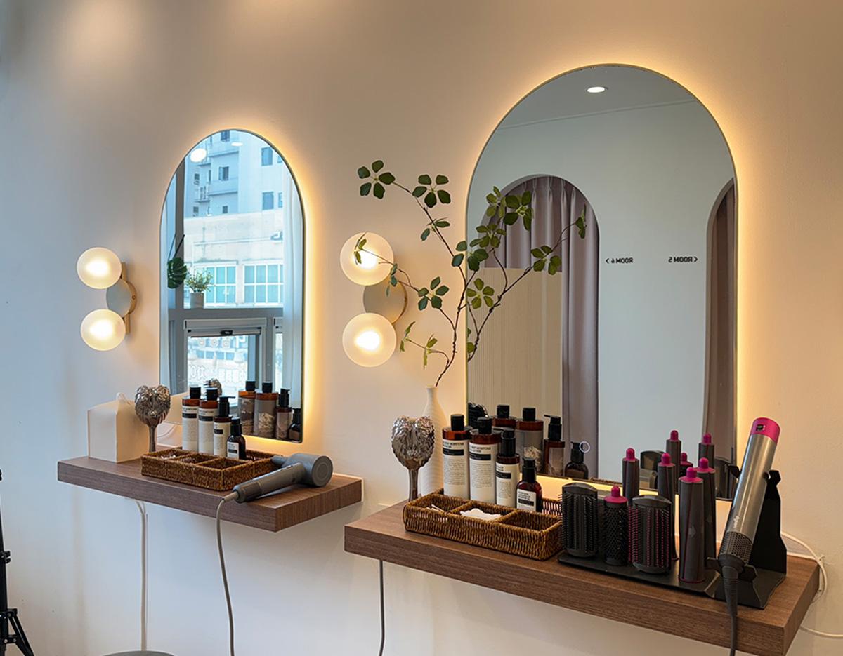 Minimal salon station with two backlit arched mirrors, wall sconces, haircare bottles, brushes, and styling tools on wooden shelves