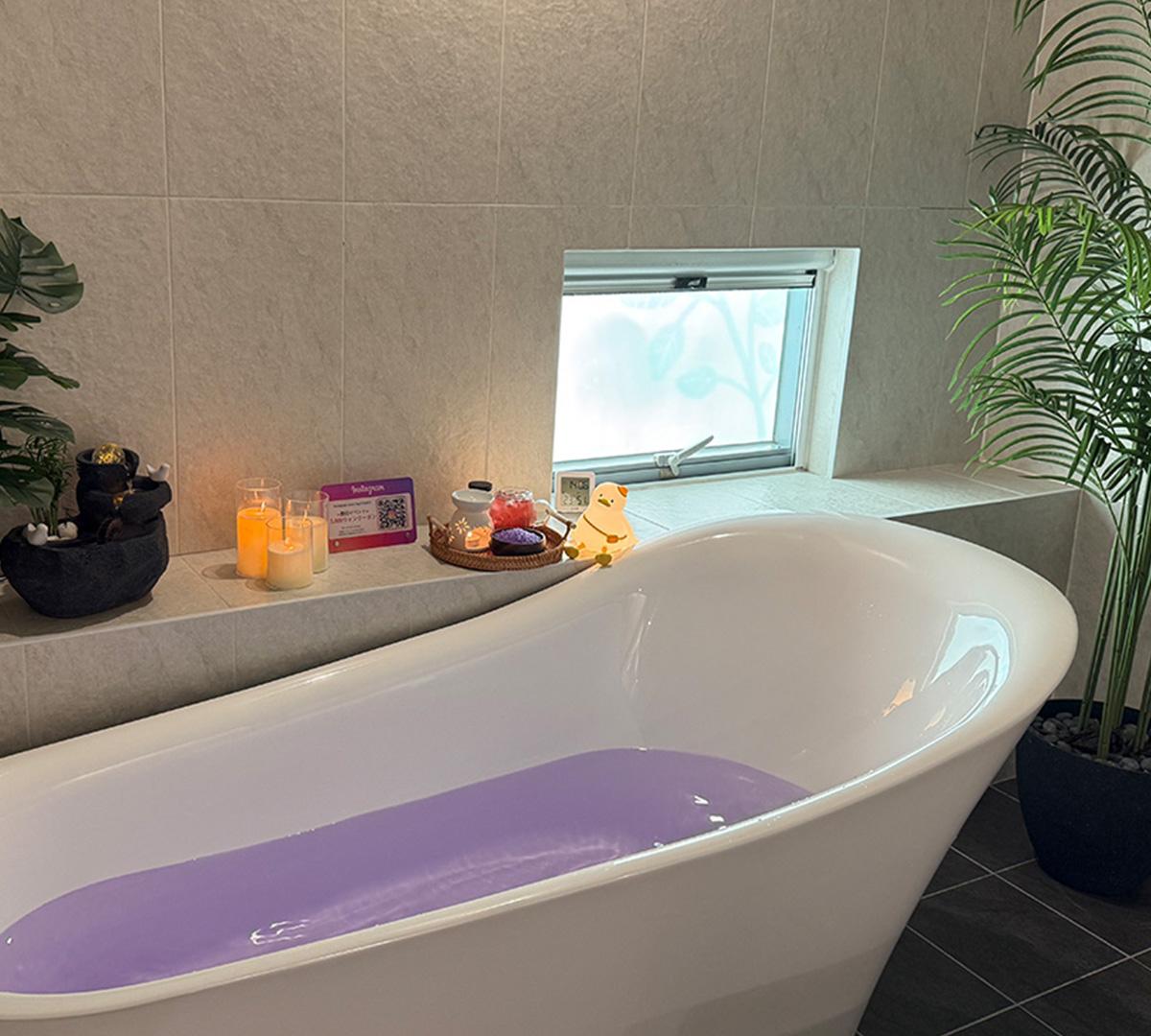 freestanding white soaking tub filled with lavender-colored bath water, candles, spa items on ledge and potted plants in private treatment room
