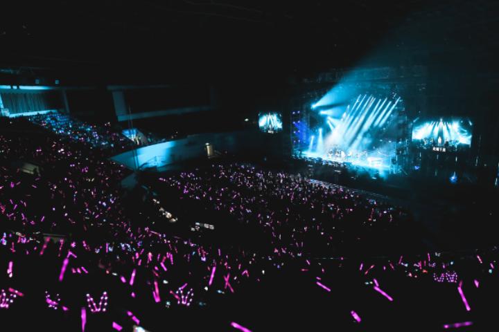 2026 KMCHART AWARDS arena packed with crowd waving purple lightsticks toward a brightly lit stage with blue spotlights.