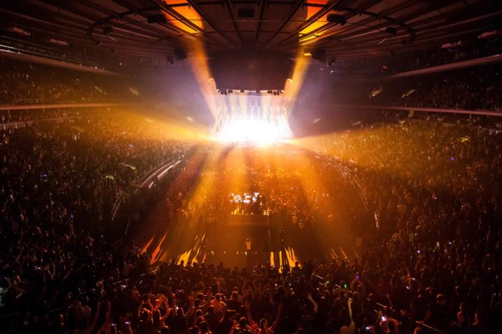 Packed arena with bright golden stage lights radiating over a standing crowd at the 2026 KMCHART AWARDS concert venue