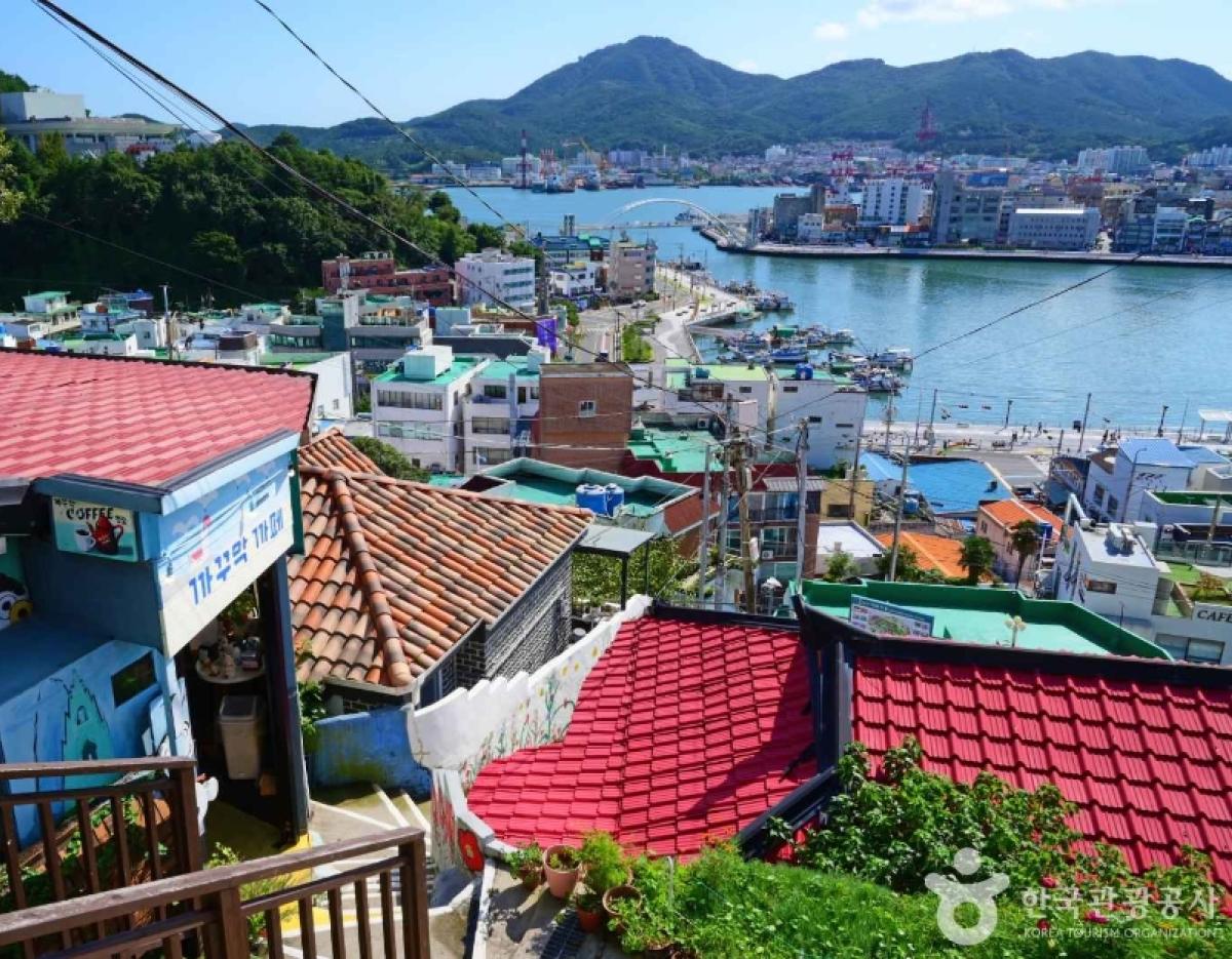 Colorful hillside village with red-tiled roofs overlooking a bay, fishing boats, bridge and distant green mountains