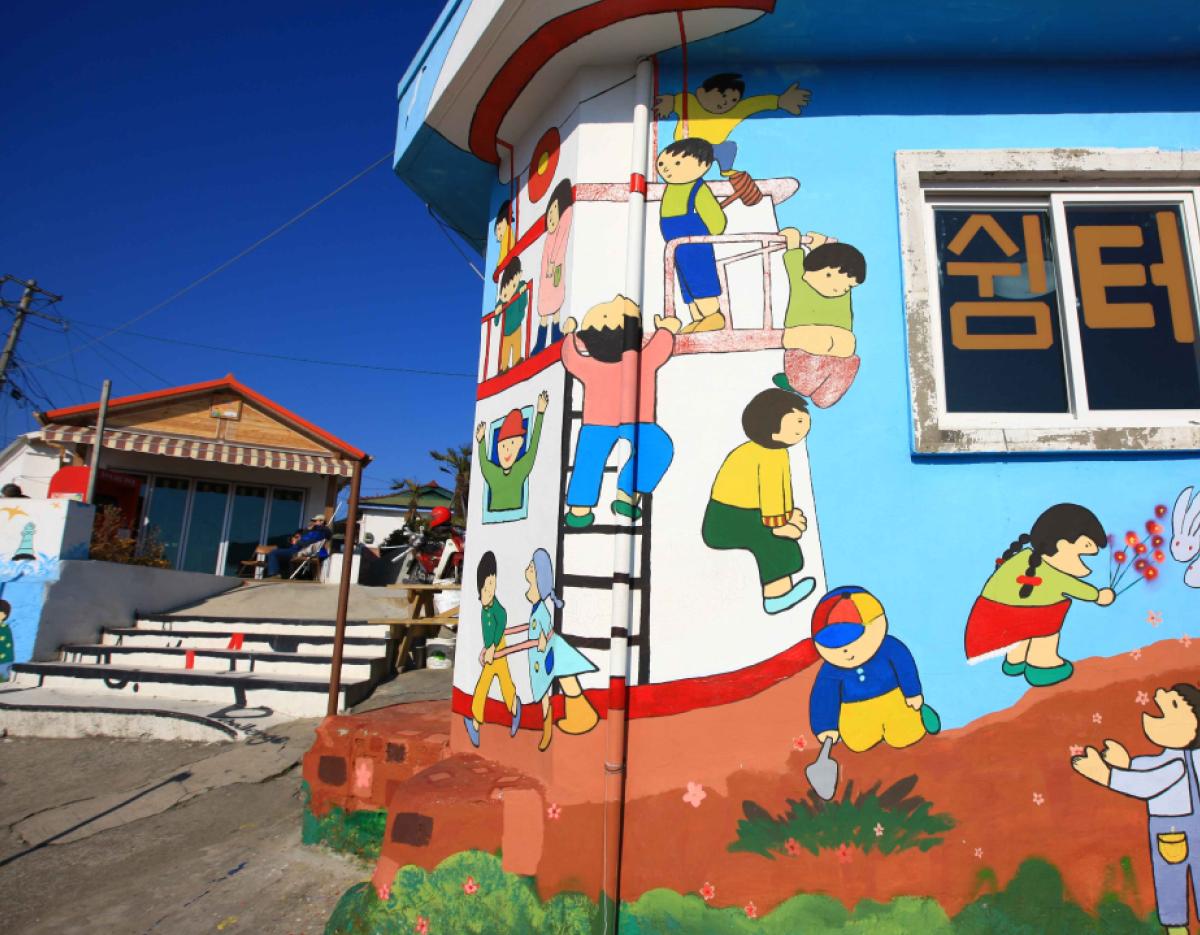 Bright blue building corner painted with colorful cartoon children climbing, playing, and gardening beside a window with Korean text