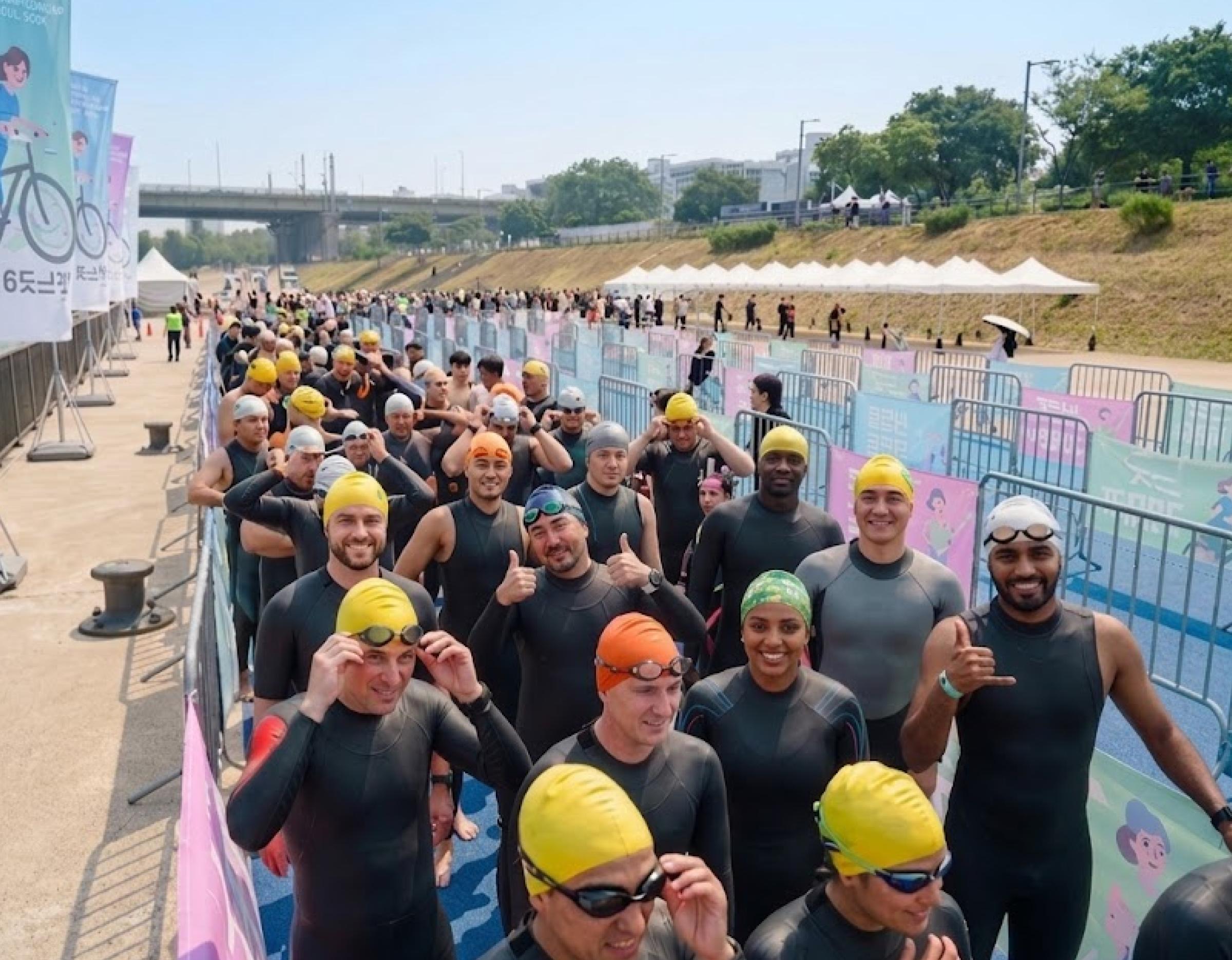 Crowd of wetsuit-clad swimmers wearing colorful swim caps queued in fenced staging area along the Han River event path