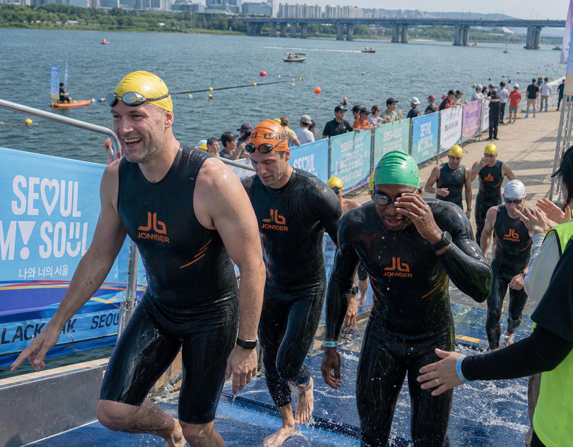Swimmers in black wetsuits and colored swim caps climbing a ramp from the Han River during a 1km race event.