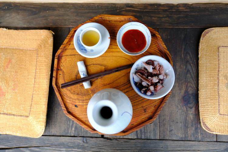 wooden tray with small ceramic soju bottle, two cups (one with amber liquor, one red sauce), bowl of sliced seafood and chopsticks