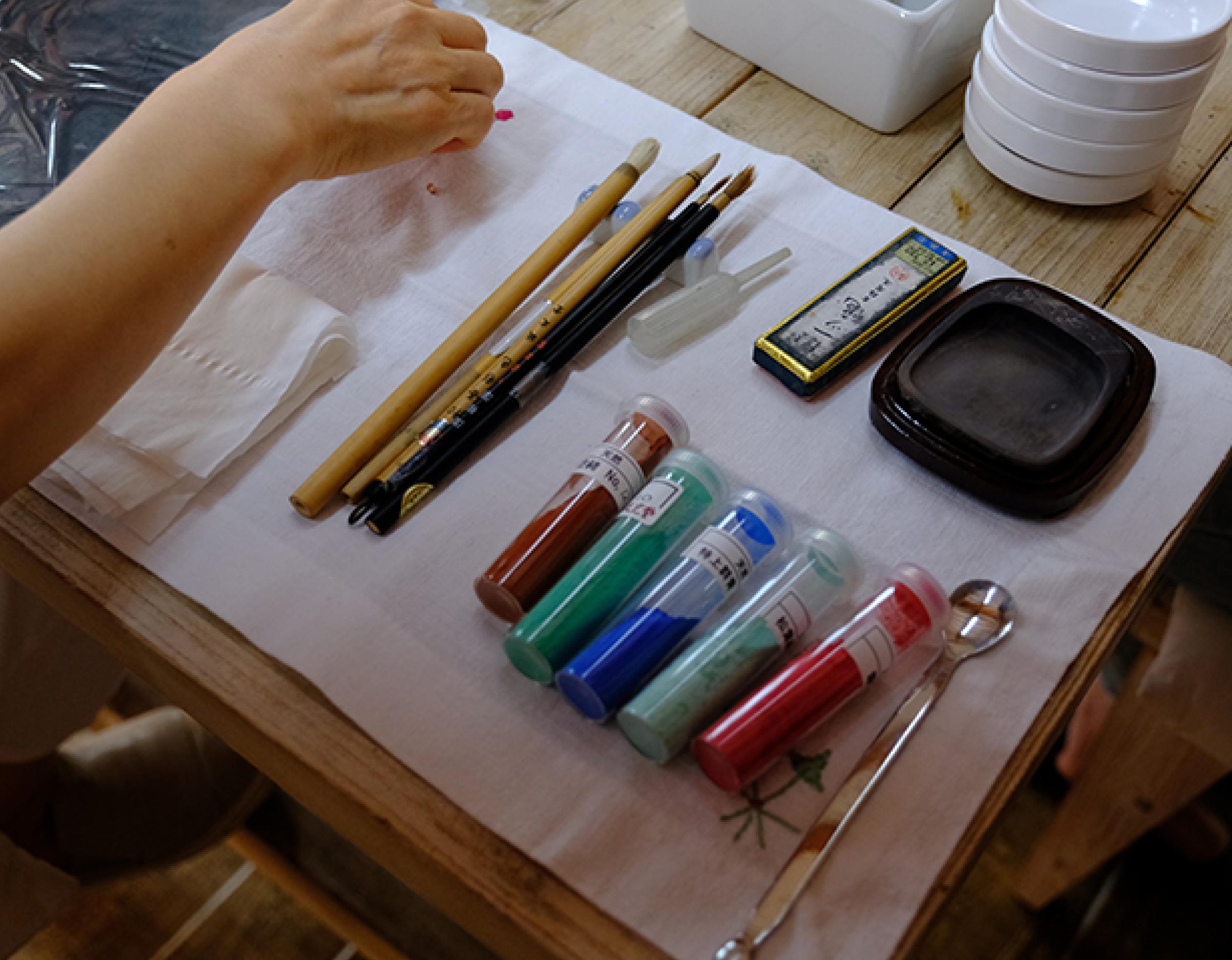 tabletop setup for Silla folk-painting fan workshop in Gyeongju: brushes, inkstone, pigment tubes, spoon and participant hand