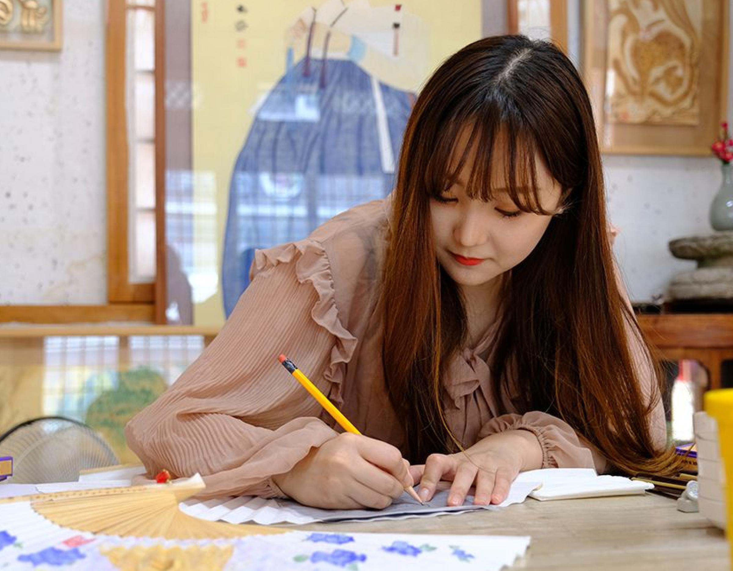 Woman in a ruffled blouse drawing traditional Silla-style folk painting on a folding fan with pencil at workshop table