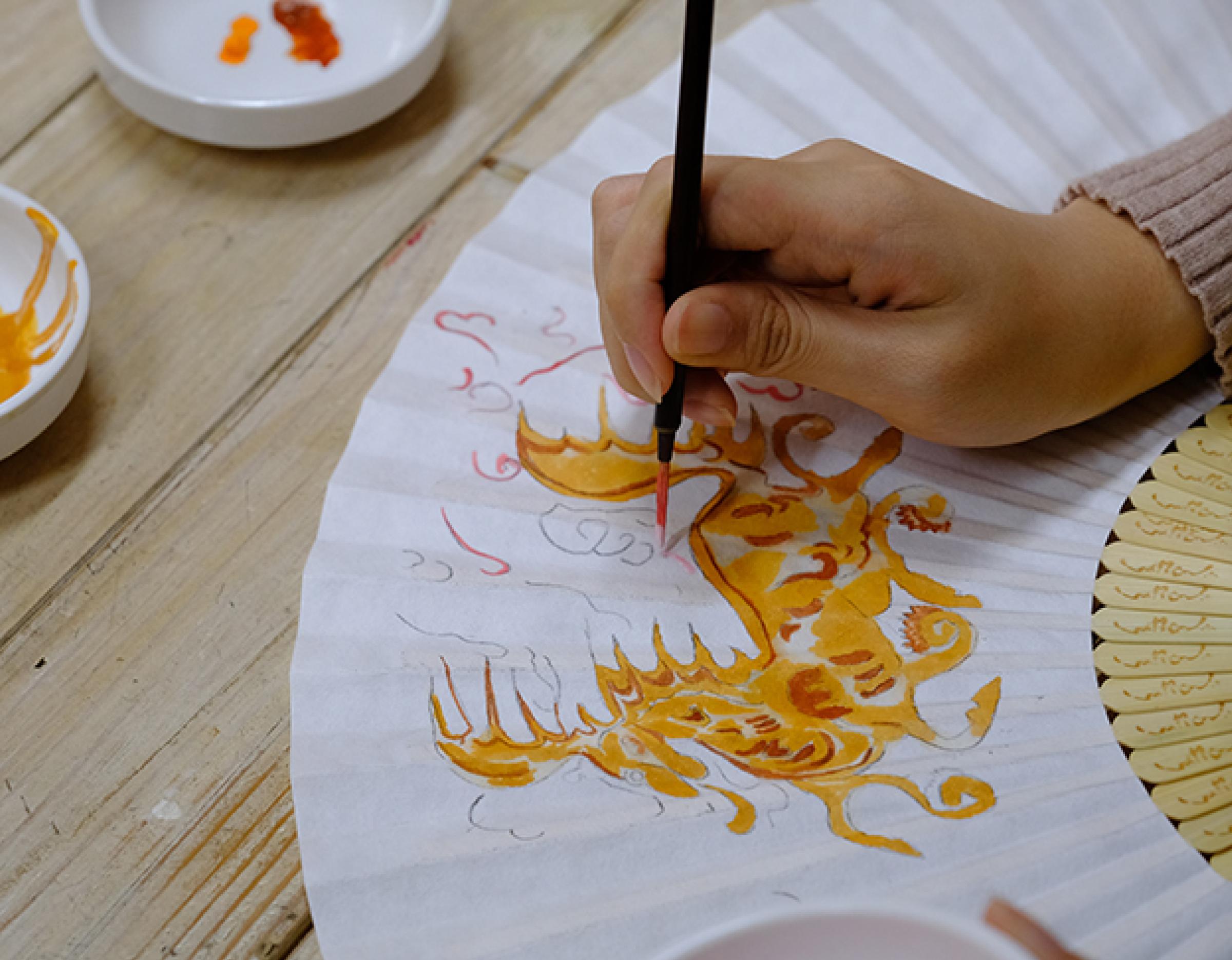 Hand painting a yellow-orange Silla-era folk motif onto a folding fan with small dish palettes nearby