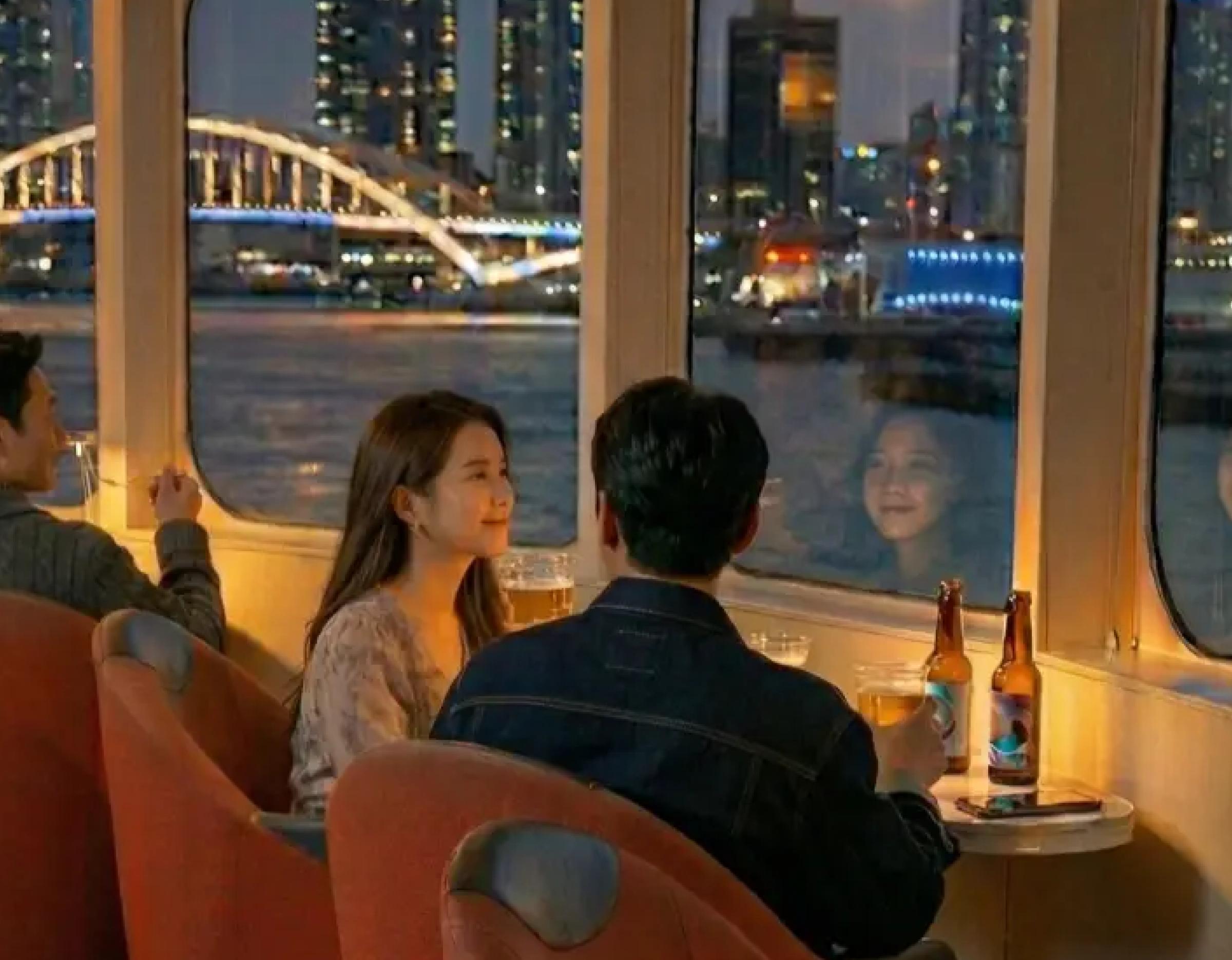 Couple inside the 부산 502인승 프리미엄 선셋/불꽃 축제 슈퍼 요트 투어 lounge sipping beer by window with lit bridge view.