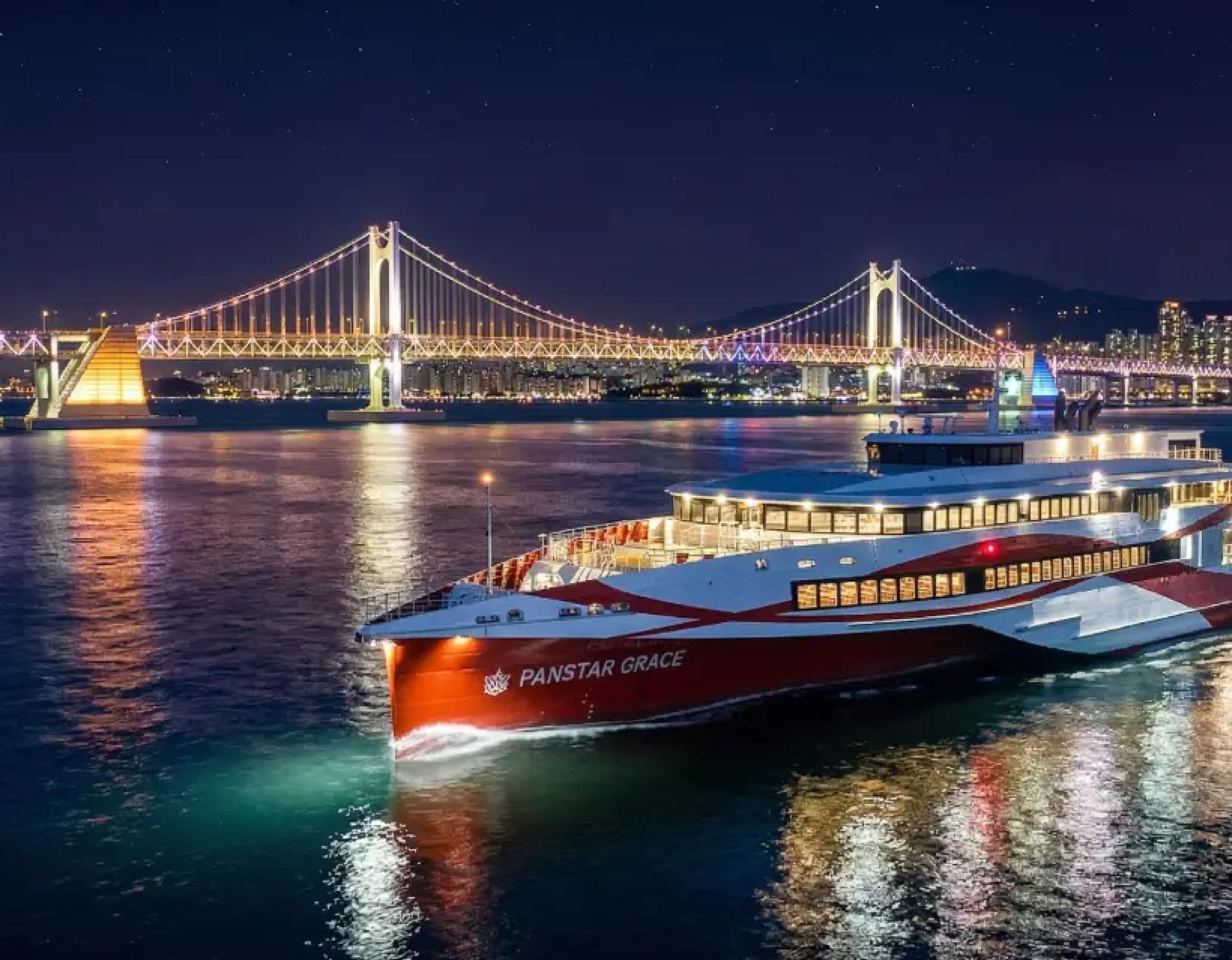 Red-and-white 502-passenger superyacht "PANSTAR GRACE" cruising at night with illuminated Gwangan Bridge and city lights reflected on water