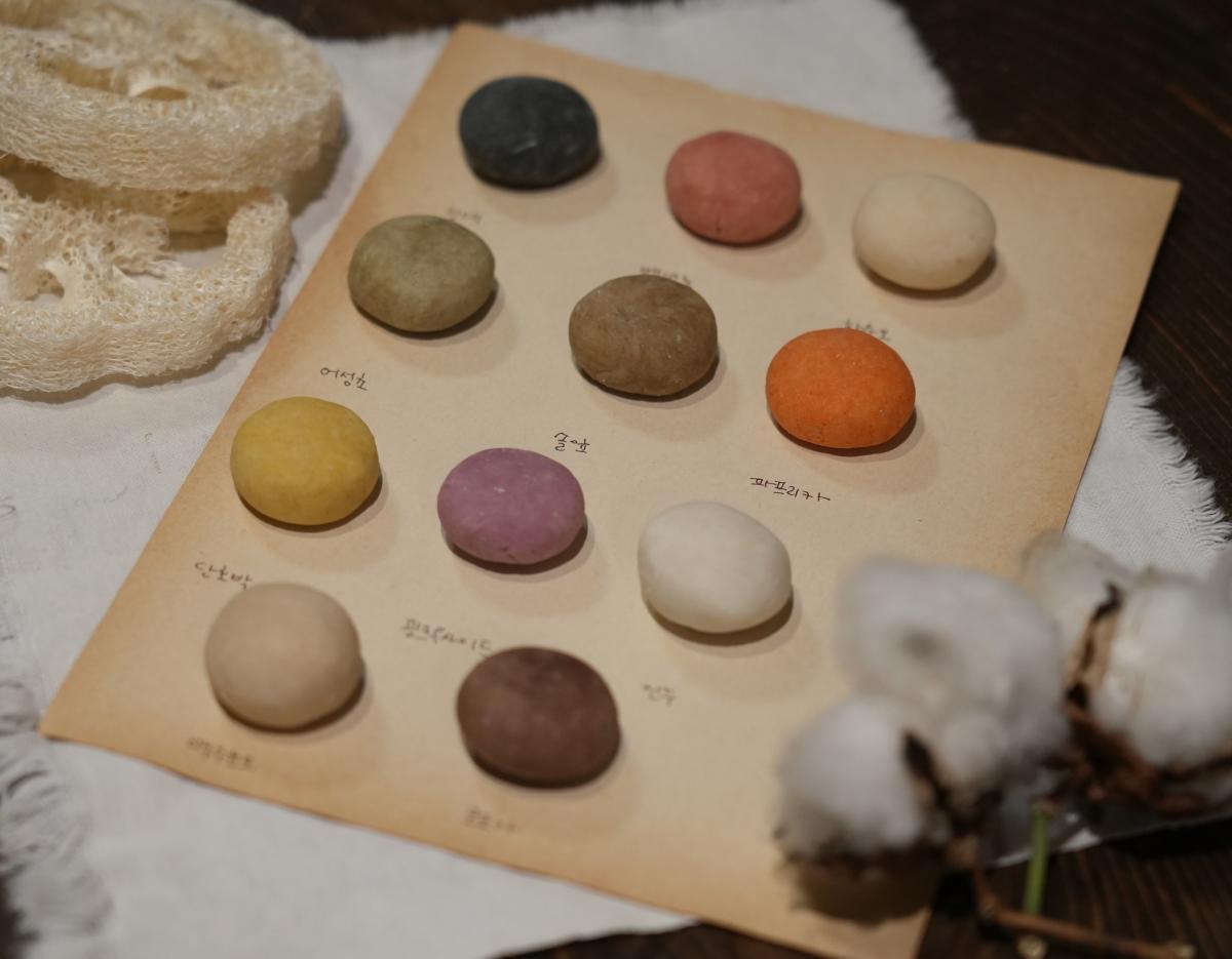 Rowed colorful round natural shampoo bars on paper labeled with Korean ingredient names, cotton bolls and loofah beside them