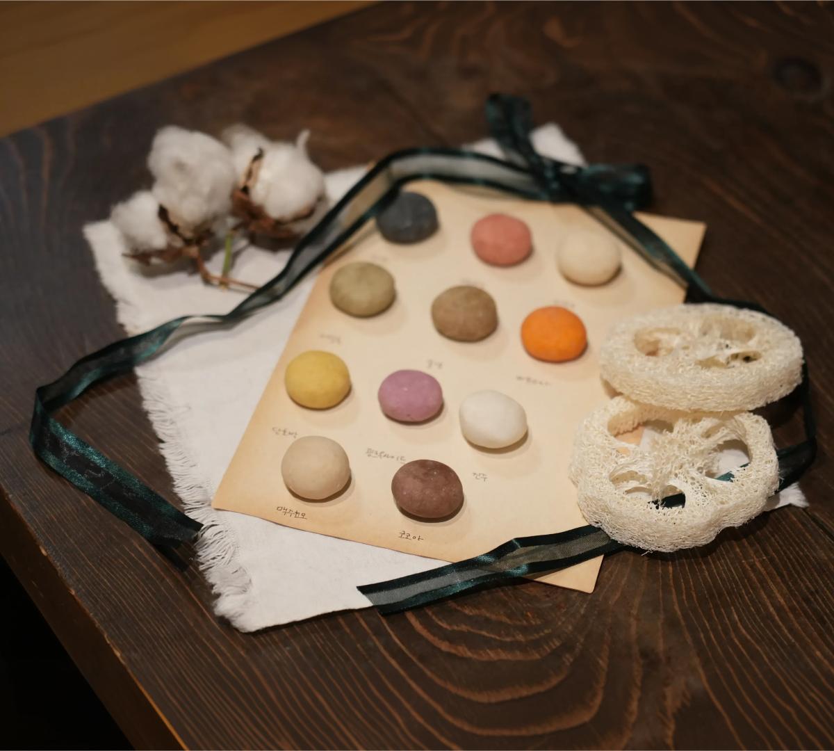 round handmade shampoo bars in assorted colors displayed on paper with loofah slices, cotton boll and dark ribbon on wooden table