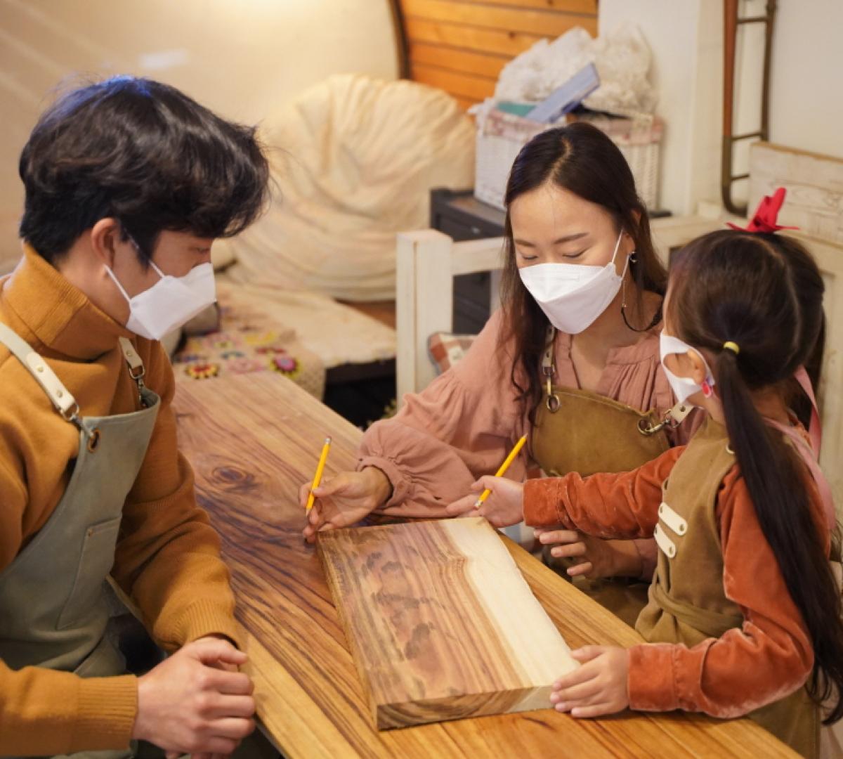 Family wearing aprons and masks sketching a wooden cutting board on a workshop table during 나무 도마 만들기 체험 | 경주