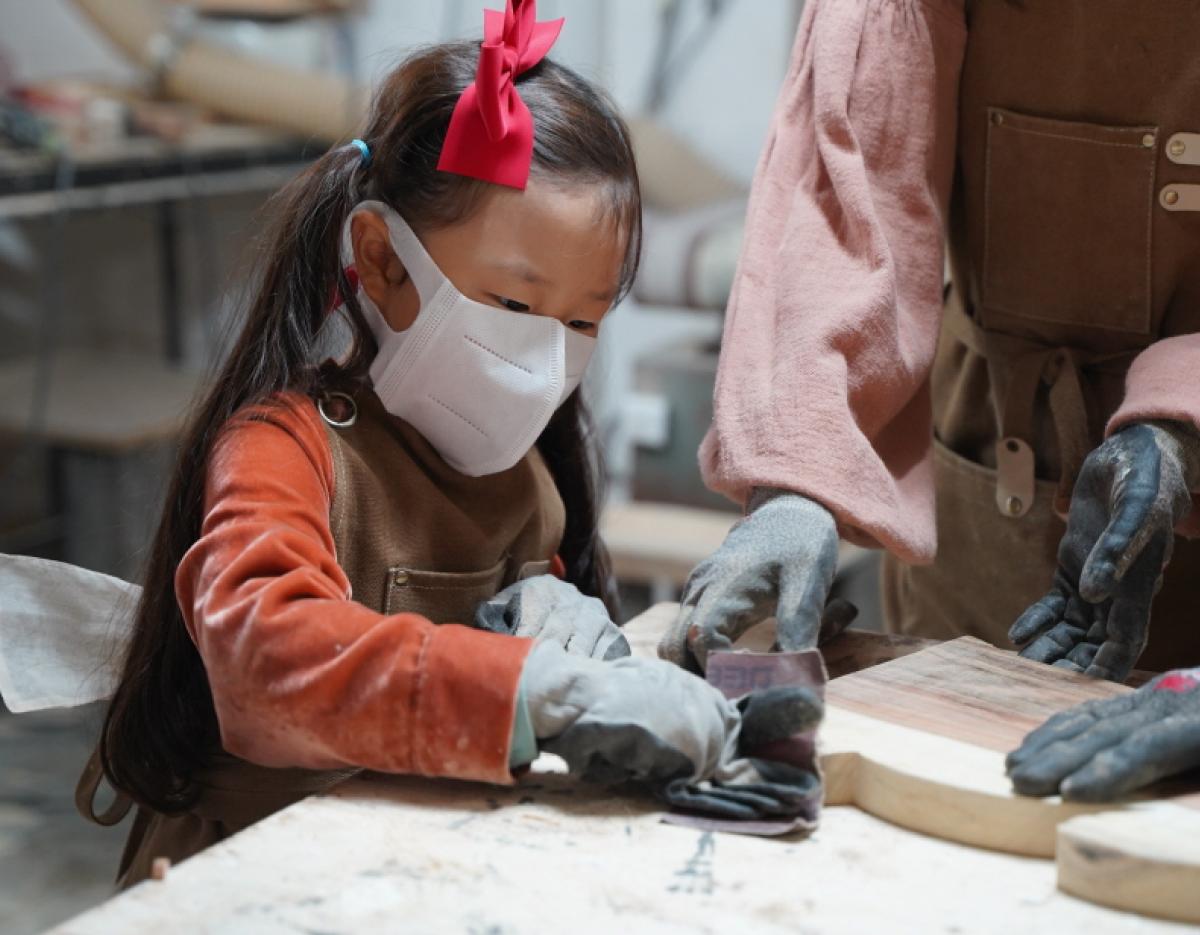 young child wearing gloves and apron sanding a wooden cutting board during 나무 도마 만들기 체험 in Gyeongju