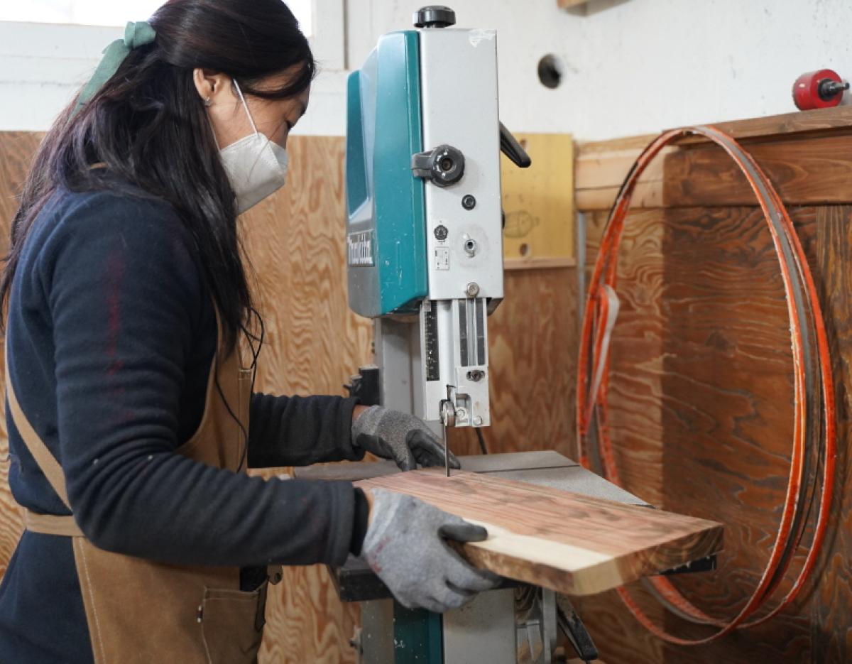 Woman wearing apron and gloves guiding wooden cutting board through band saw during 나무 도마 만들기 체험 | 경주 workshop