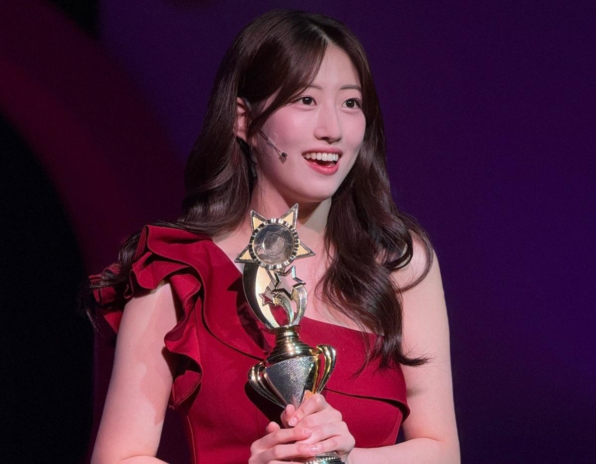 woman in a red ruffled one-shoulder dress holding a gold star-topped trophy against a purple stage background