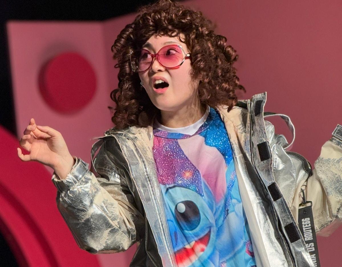 actor in curly wig and metallic silver jacket gesturing on stage, wearing colorful graphic tee with cartoon blue character
