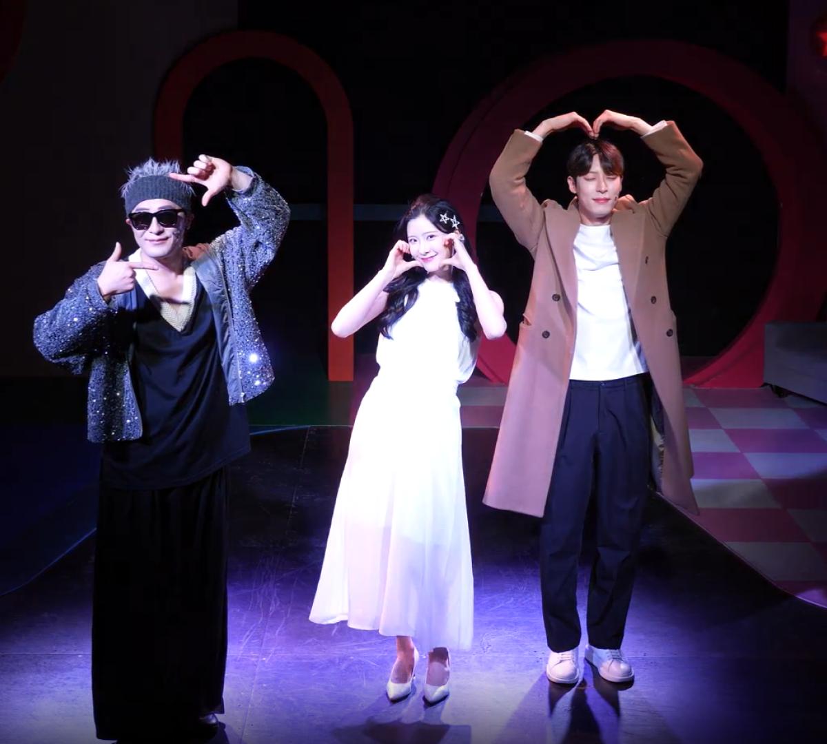 Three stage performers under spotlight: left in sparkly jacket, center woman in white dress, right man in long coat making a heart gesture.