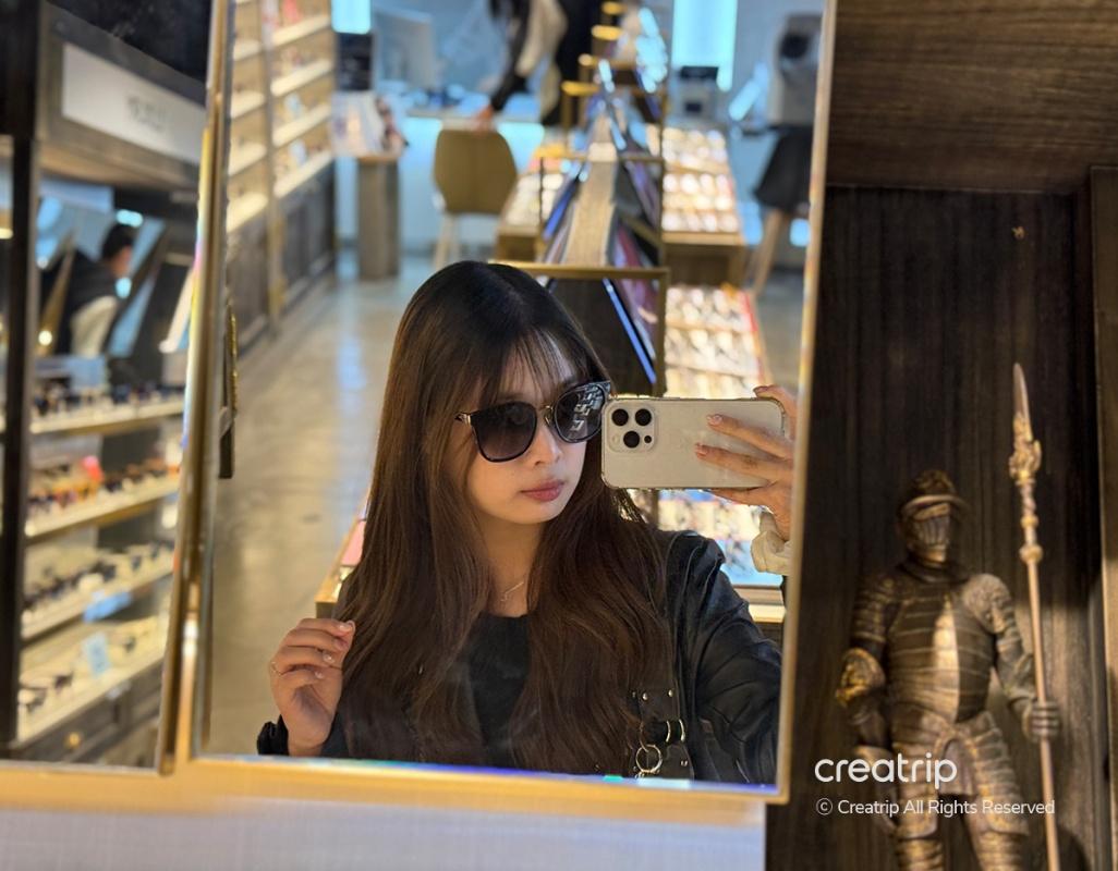 woman with long dark hair taking mirror selfie inside boutique eyewear shop, glasses displays and armor statue visible