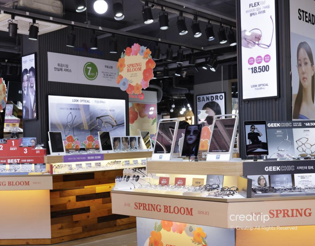 Bright Look Optical store interior with display tables of K-pop style eyeglasses, screens, signage reading "SPRING BLOOM" and price tags