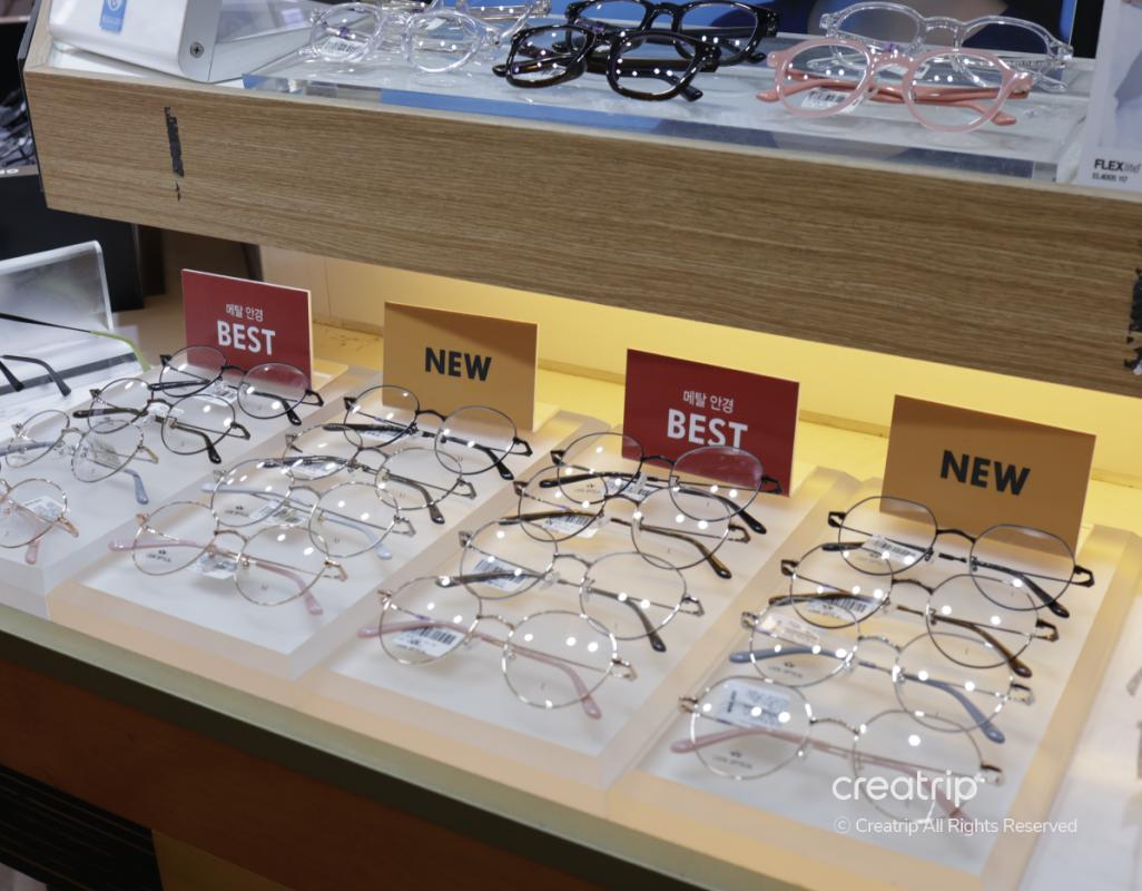 Display of multiple round and rectangular prescription eyeglass frames on lit trays labeled "NEW" and "BEST" at 룩옵티컬 강남역점.