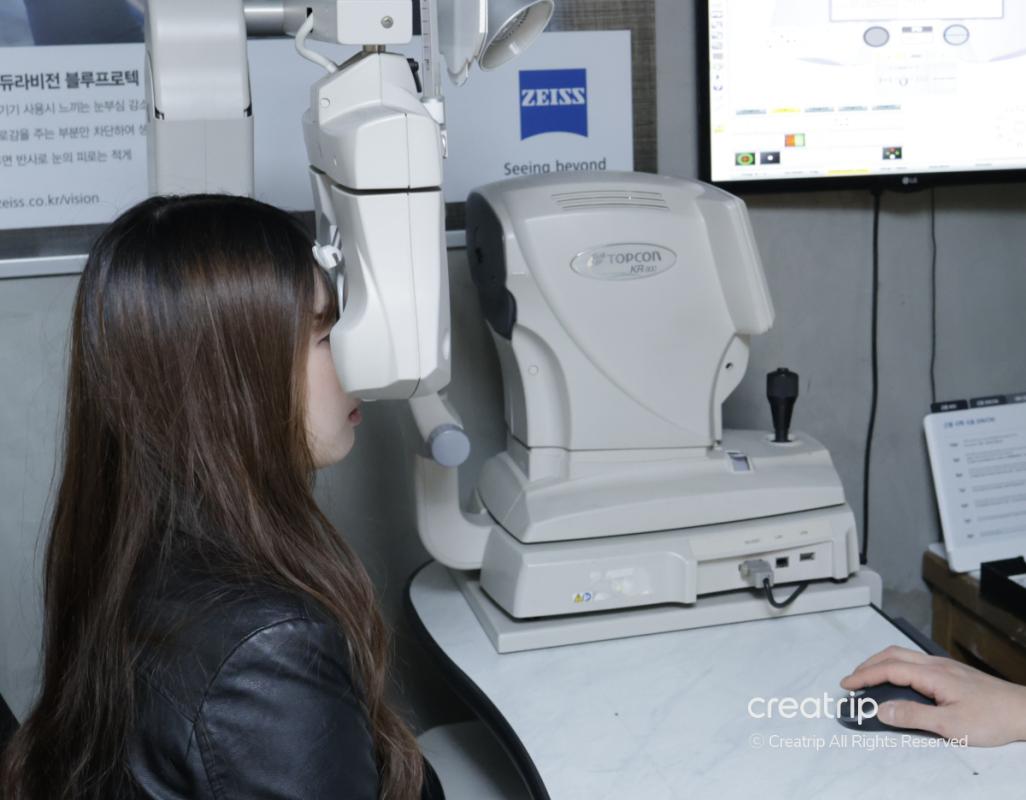 woman seated at an ophthalmology machine undergoing an eye exam with technician operating the device in a clinic