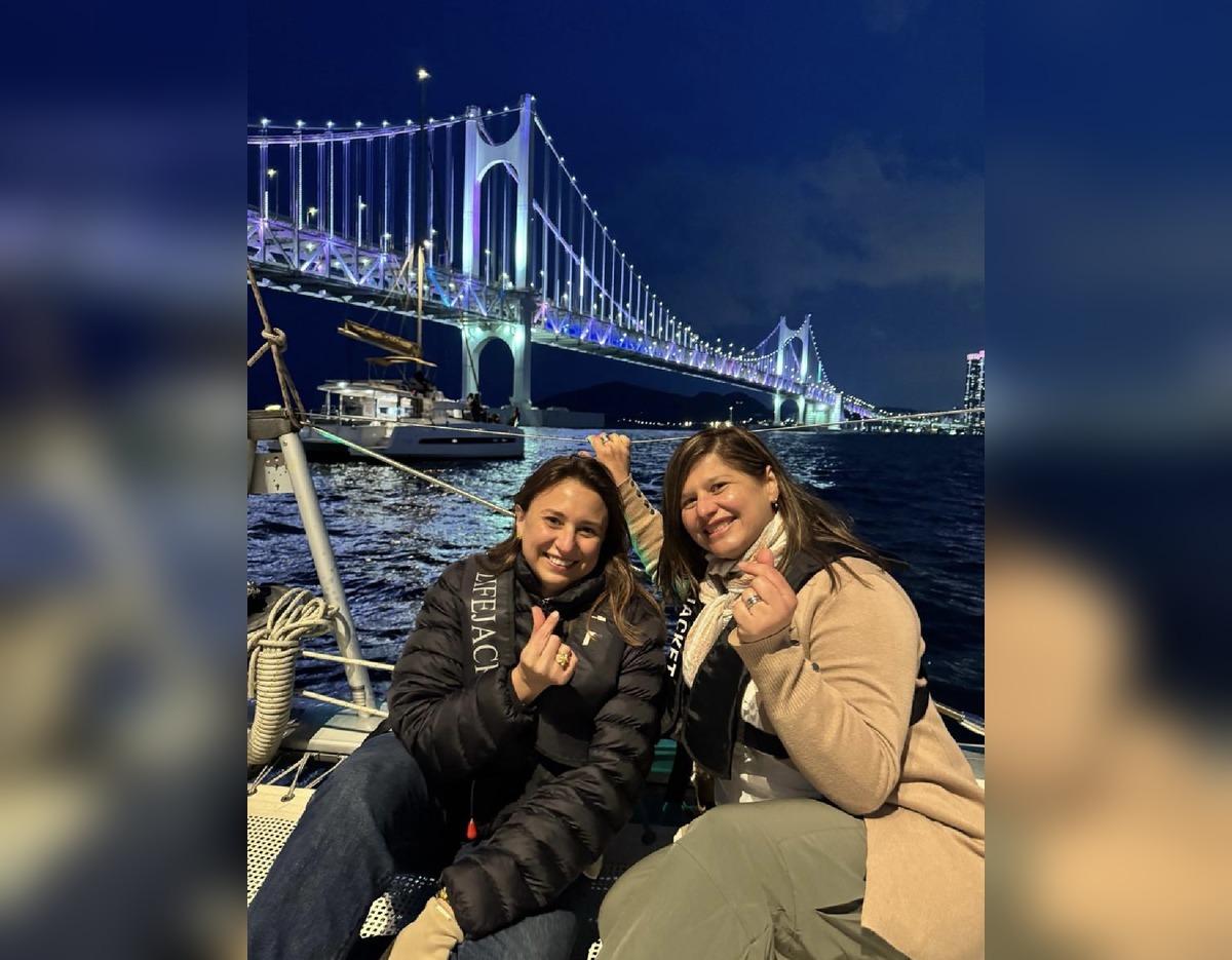 couple on a yacht at night making finger-heart gestures with illuminated Gwangan Bridge and city lights in background