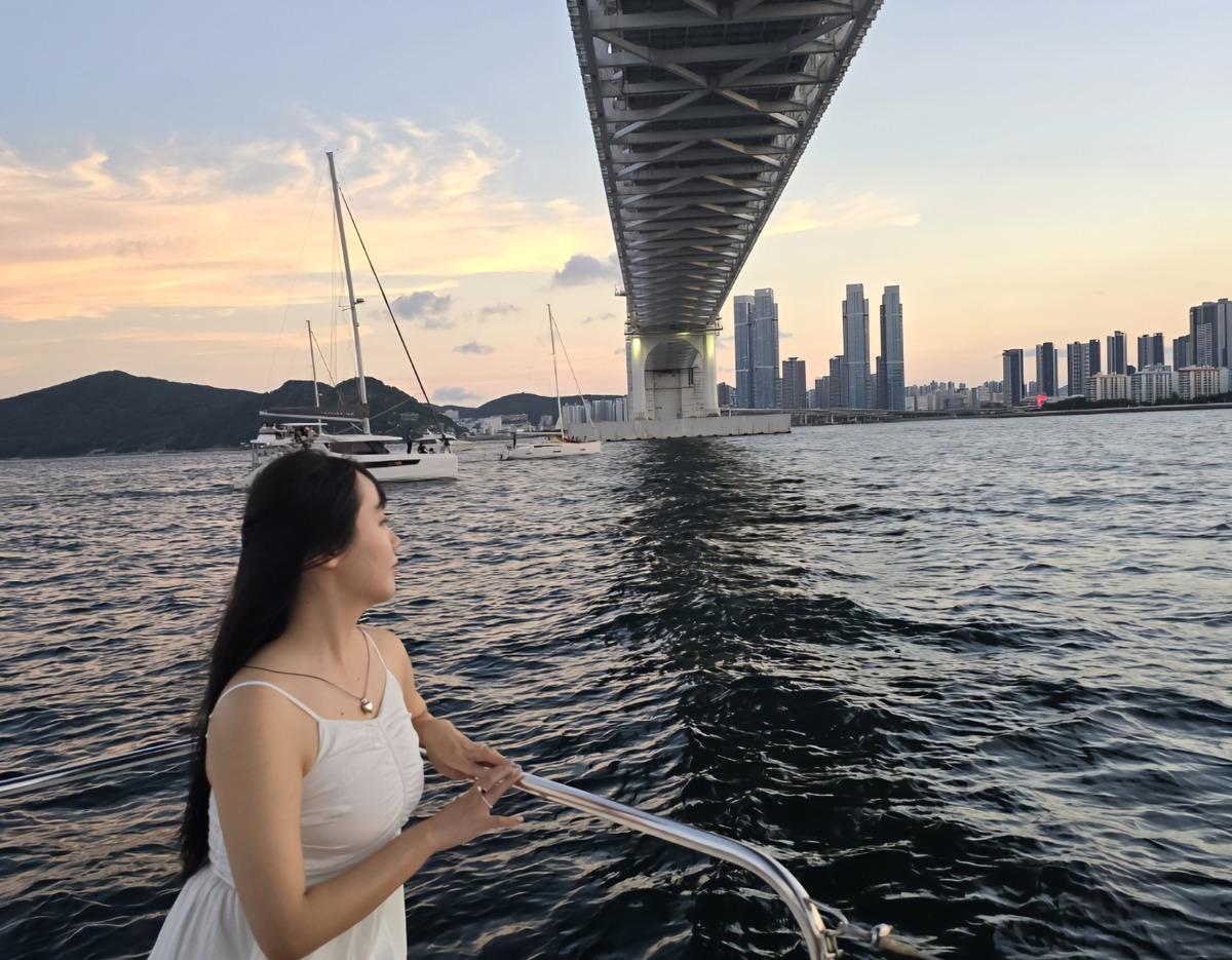 woman in white dress leaning on yacht rail under Gwangan Bridge at sunset, city skyline and sailboats in background