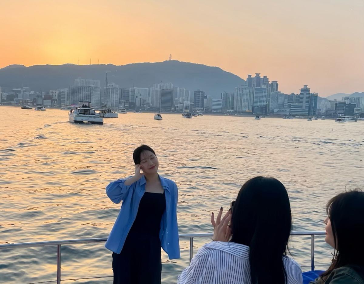 Three passengers on a yacht railing at sunset, woman posing for photo with Busan skyline and calm harbor waters behind.