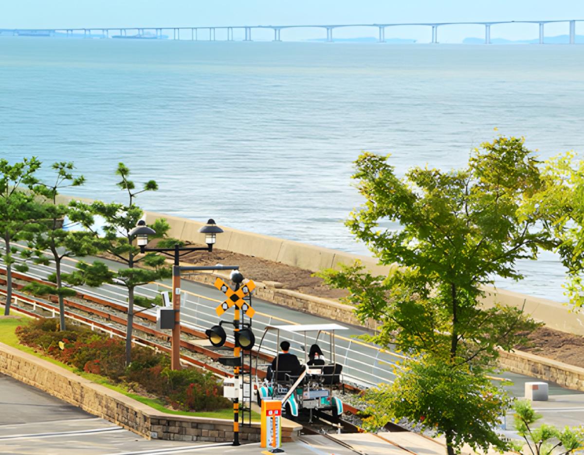 seaside rail bike with two riders on tracks, coastal path, trees in foreground and Incheon Bridge across the sea background