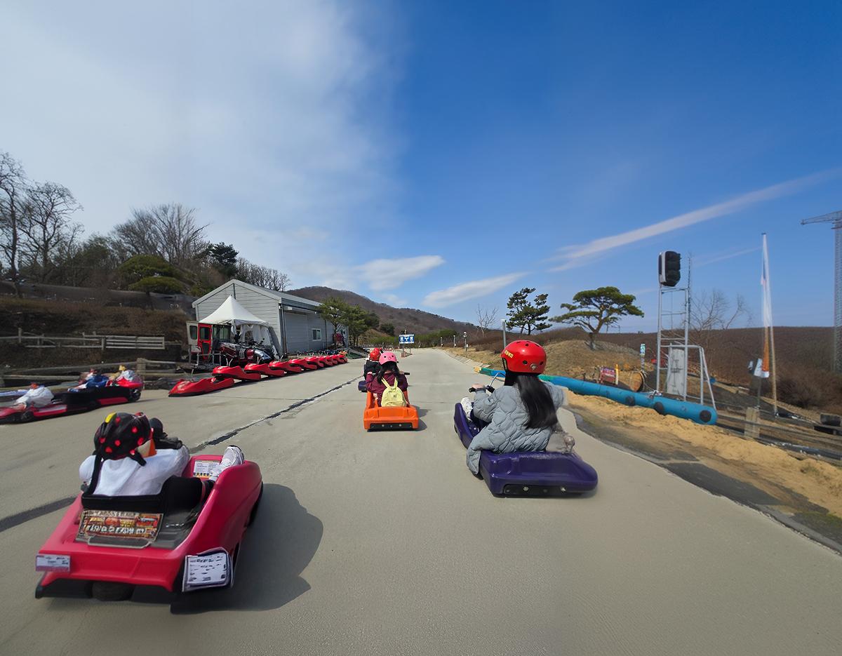 Riders in colorful helmets on single-seat luge carts racing down a paved coastal track at Ganghwa luge facility
