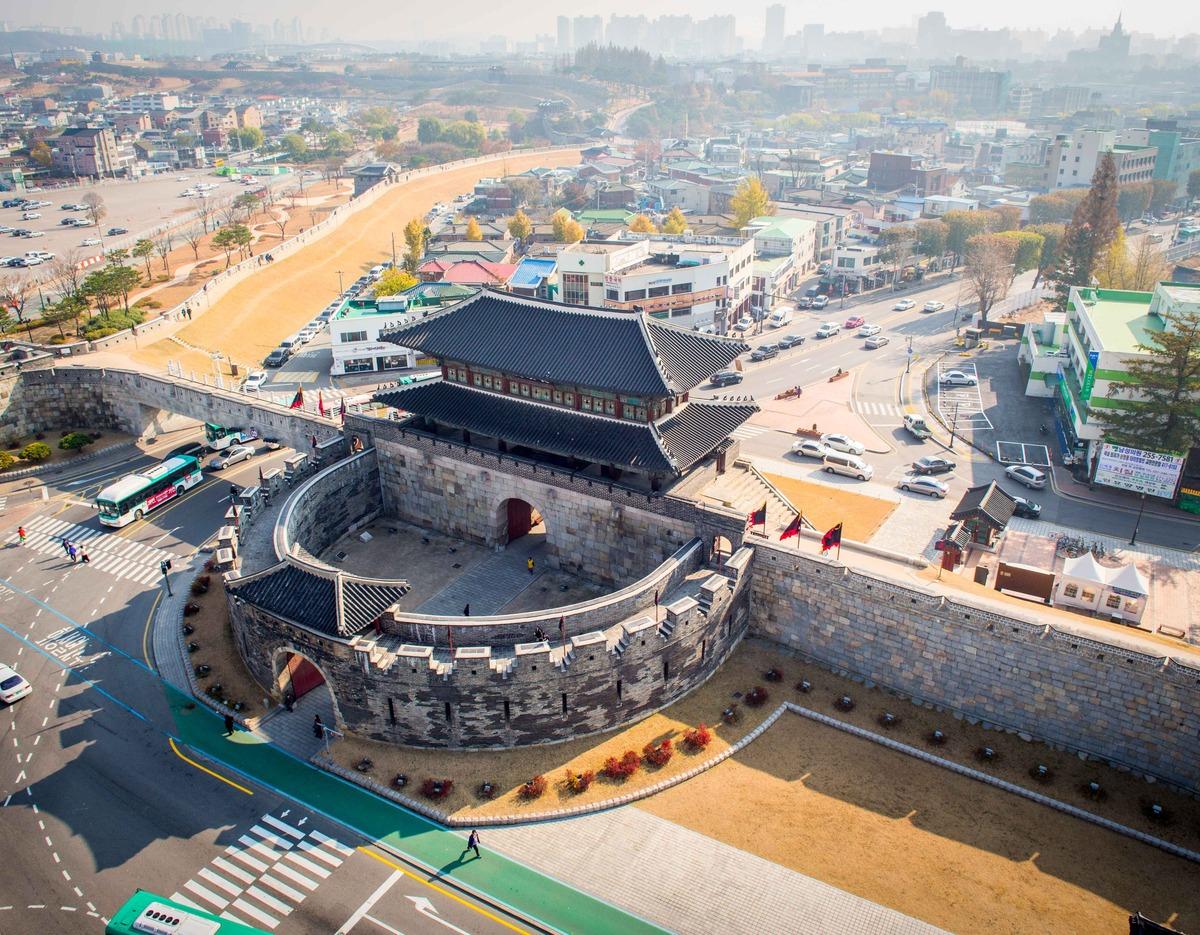 Aerial view of Suwon Hwaseong Fortress gate with curved stone walls, tiled pavilion and surrounding city streets.