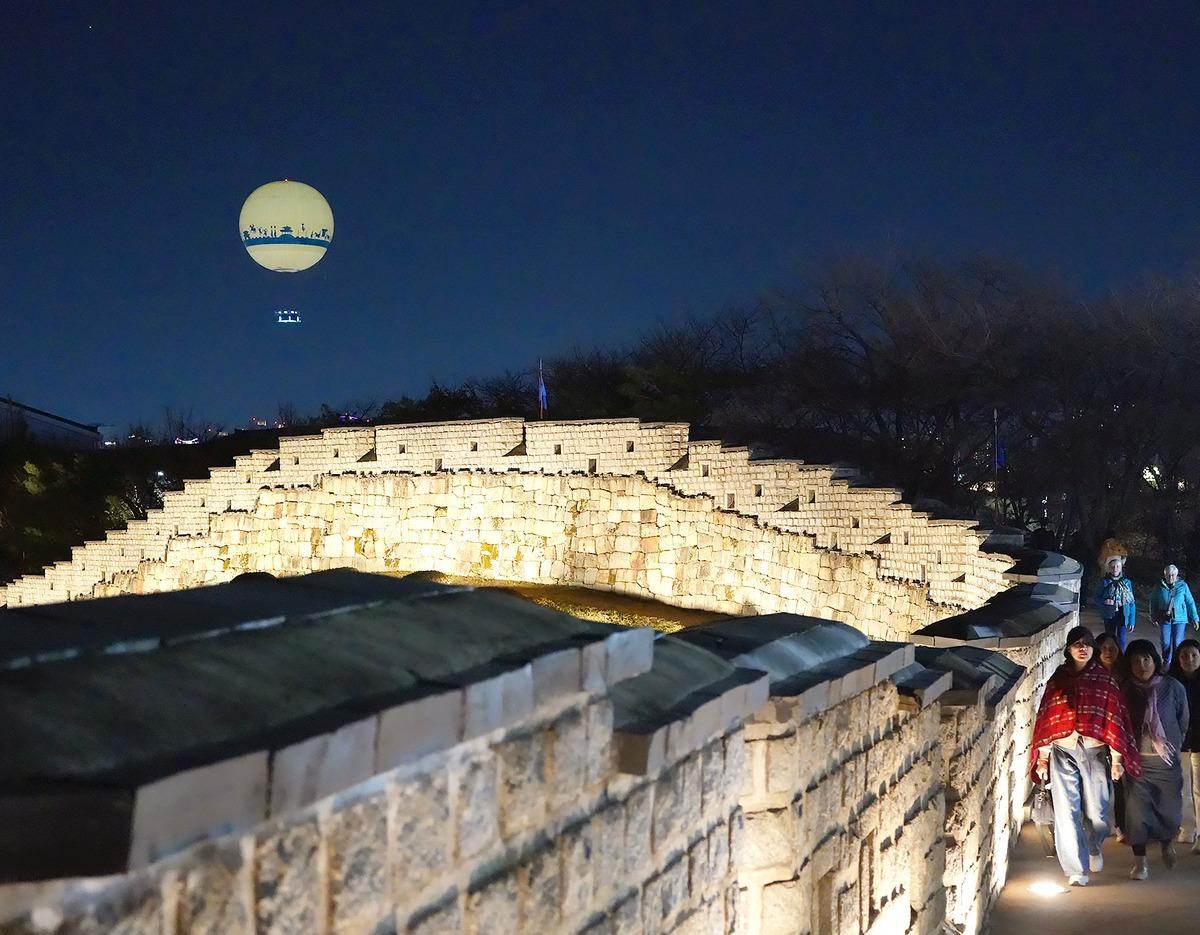 Illuminated Hwaseong Fortress stone wall at night with visitors walking along the lit path and a glowing tethered balloon above