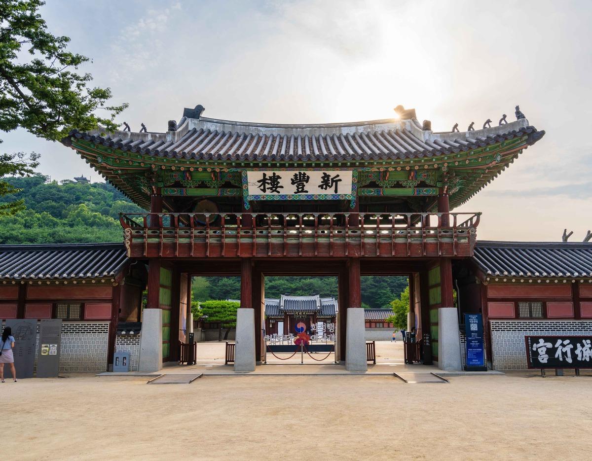 Hwaseong Haenggung Palace wooden gate with ornate painted eaves opening to courtyard, hills and sunlit sky behind