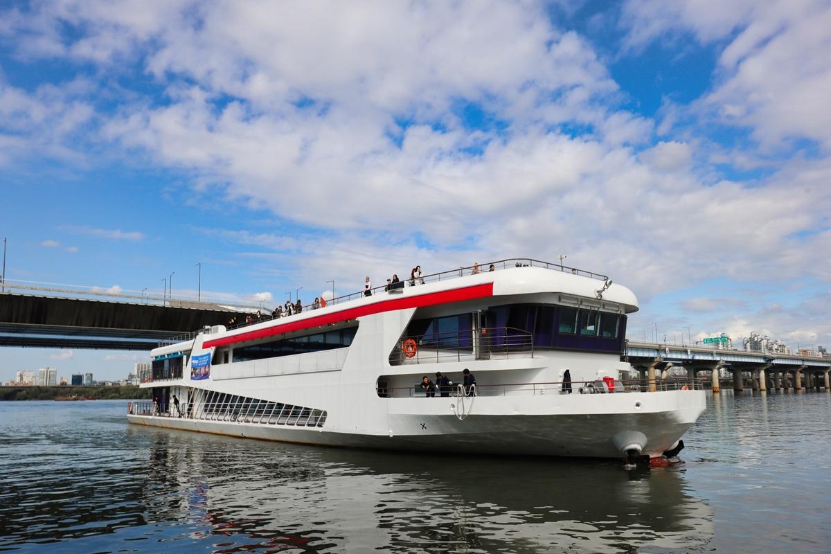 White multi-deck sightseeing cruise boat with red stripe cruising on Han River near Yeouido bridges under blue sky