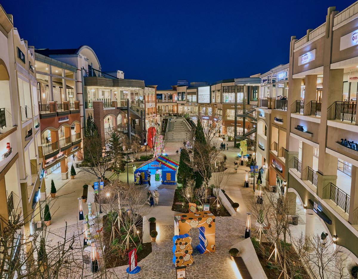 Outdoor nighttime view of Paju Premium Outlets open-air mall with illuminated stores, central walkway and colorful installations