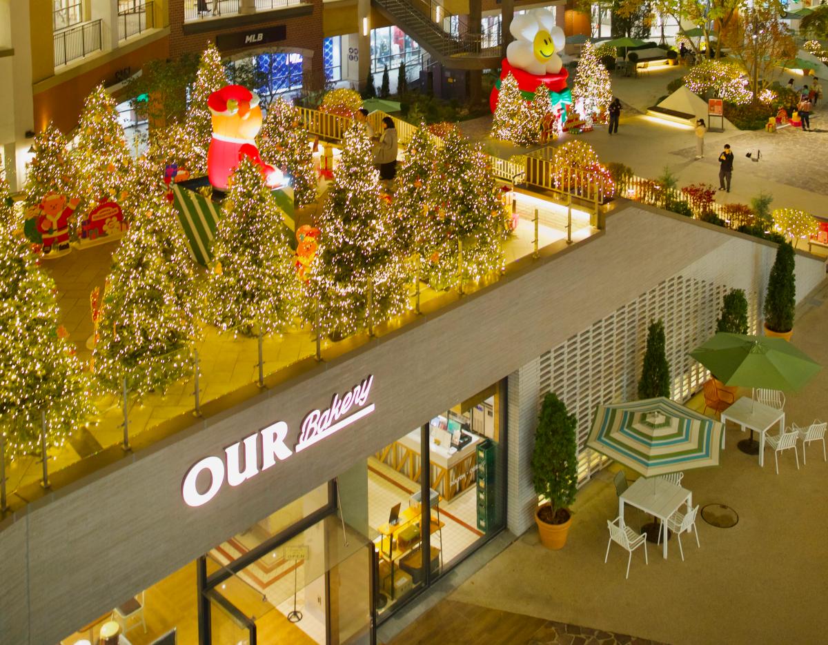 Outdoor mall scene at Paju Outlet shuttle stop with lit Christmas trees above storefront "OUR Bakery" and terrace seating.