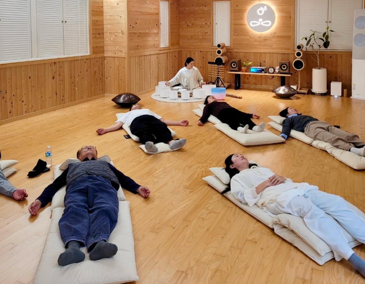 Wood-paneled meditation room with participants lying on floor cushions during a sound bath and tea-centered wellness session.