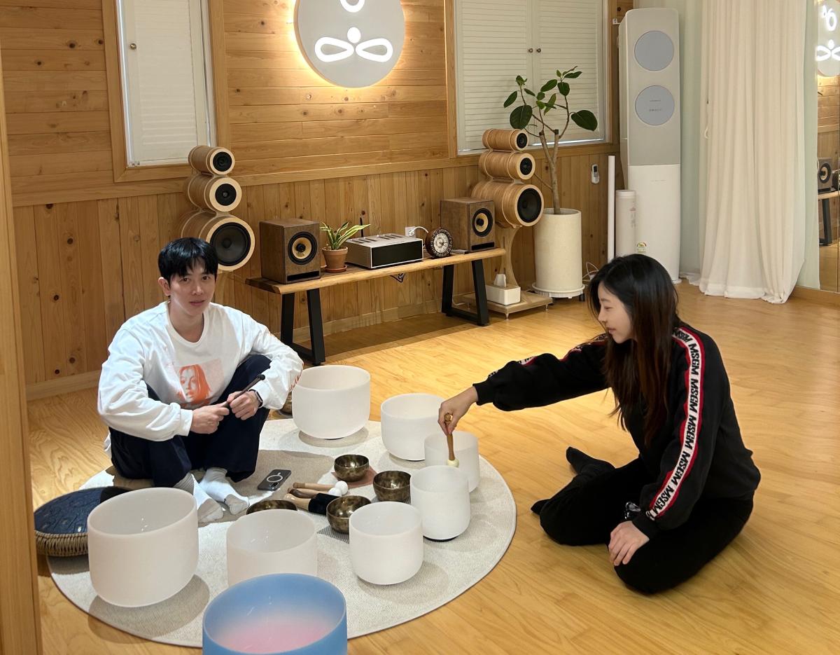 Two people sitting on a wooden floor around crystal singing bowls and Tibetan bowls in a cozy cypress-scented meditation room.