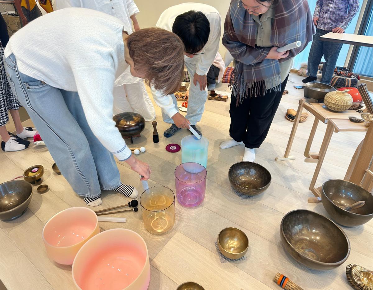 Participants kneeling around colorful crystal and metal singing bowls and handpans on a light wooden floor during a sound-bath session